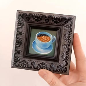 Puede incluir: Una pintura en miniatura de una taza de caf&eacute; azul con un dise&ntilde;o de arte latte, enmarcada en un marco negro adornado.