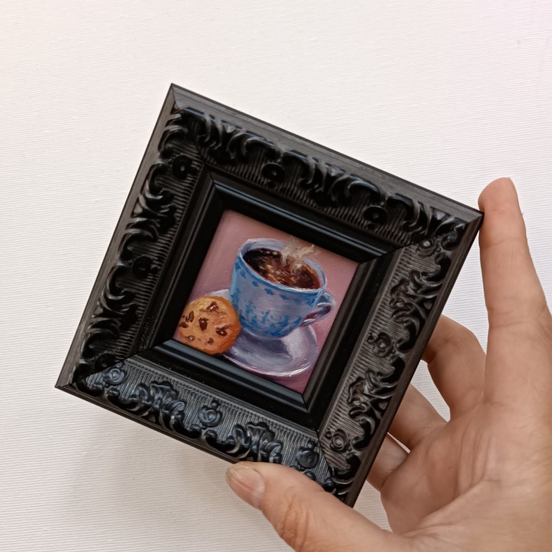 May include: A small framed painting of a blue and white teacup with steaming coffee and a chocolate chip cookie on a saucer.