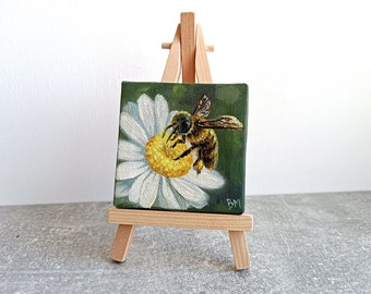 May include: A small painting on a wooden easel depicts a bee collecting pollen from a white daisy with a yellow center. The background is a dark green.