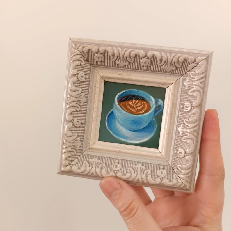 Puede incluir: Una peque&ntilde;a pintura enmarcada de una taza de caf&eacute; azul con un dise&ntilde;o de arte latte. La taza est&aacute; sobre un platillo azul y el fondo es verde.