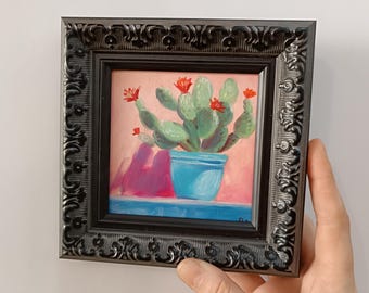 Peut inclure: Peinture à l'huile encadrée d'un cactus en pot bleu avec des fleurs rouges. Le tableau a un fond rose et une surface bleue. Le cadre noir est orné de détails.