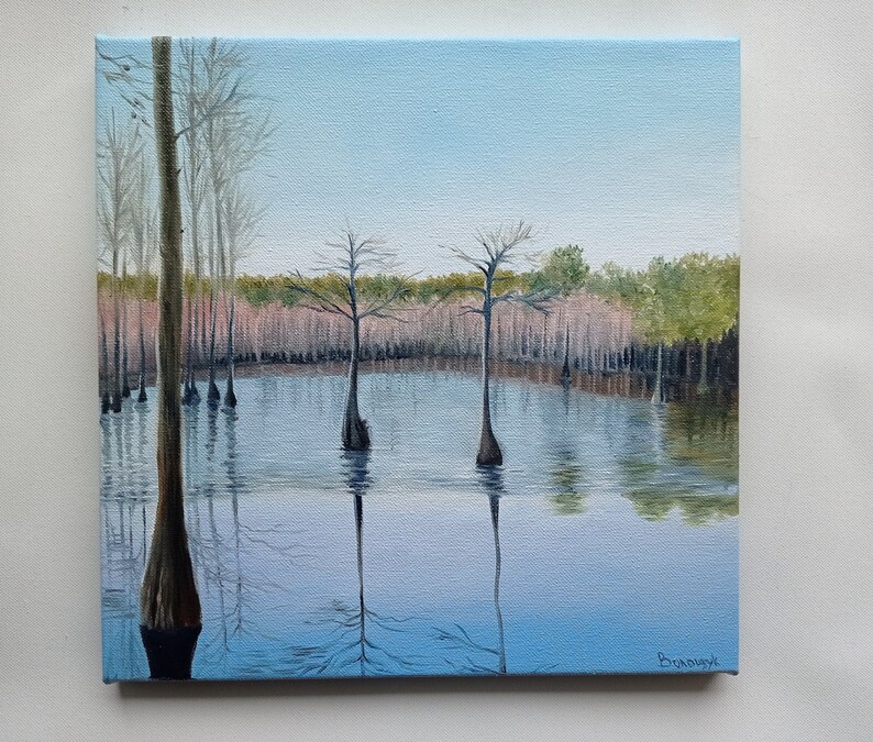 Puede incluir: Una pintura que representa una escena de pantano con tres cipreses en el agua. El agua es de color azul claro y el cielo es de color azul claro. Los &aacute;rboles est&aacute;n desnudos y el agua est&aacute; tranquila. La pintura est&aacute; firmada "Brannock" en la esquina inferior derecha.