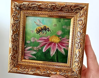 May include: A framed miniature painting of a bee hovering near a pink coneflower with a yellow center. The painting is set in a gold ornate frame. The background is green with other pink flowers.