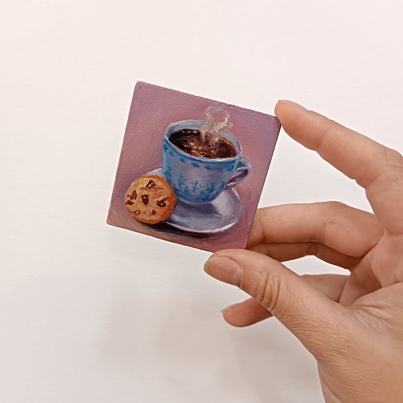 May include: A small painting of a blue and white teacup filled with coffee and steam rising from the top. A chocolate chip cookie sits on a saucer next to the teacup. The painting is on a pink background.
