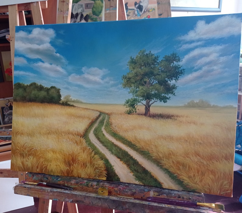 Puede incluir: Una pintura de un camino de tierra que serpentea a trav&eacute;s de un campo de trigo dorado. Un solo &aacute;rbol se alza en medio del campo, con un cielo azul y nubes blancas en el fondo.