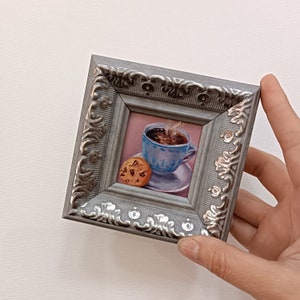 May include: A small framed painting of a cup of coffee with steam rising from it and a chocolate chip cookie on a saucer. The frame is silver with an ornate design.