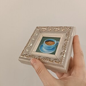 Puede incluir: Una peque&ntilde;a pintura enmarcada de una taza de caf&eacute; con arte latte. El marco es blanco con un dise&ntilde;o ornamentado de color plateado.