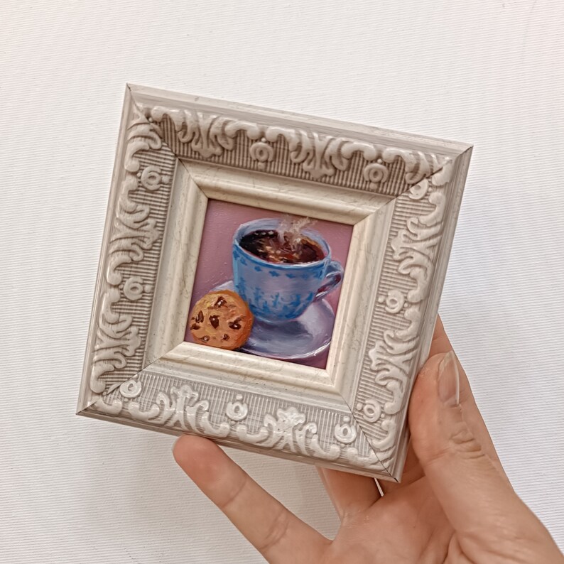 May include: A framed painting of a cup of coffee with steam rising from it and a chocolate chip cookie on a pink background.