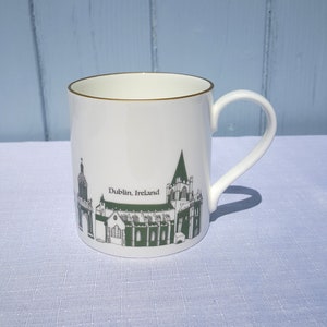 Può includere: Tazza in ceramica bianca con bordo dorato e manico curvo. La tazza presenta un'illustrazione verde e bianca di una chiesa con la scritta "Dublin, Ireland". La tazza è appoggiata su una superficie azzurra.