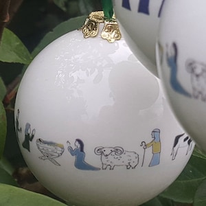 以下が含まれることがあります： 金色の縁取りが施された白い磁器製のクリスマスオーナメントが2つ。1つのオーナメントには、飼い葉桶、羊、2人の人物が描かれたクリスマスの場面が描かれている。もう1つのオーナメントには、青いローブを着た2人の人物が描かれている。