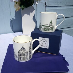 May include: Two white ceramic mugs with gold trim. One mug features a black and white illustration of Dublin, Ireland, with the text "Dublin, Ireland". The other mug features a black and white illustration of the General Post Office in Dublin, Ireland, with the text "Mandy Bullivant". Both mugs are sitting on a blue box with the text "English Fine Bone China".