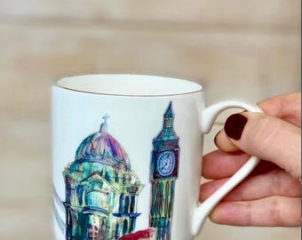 May include: White ceramic mug featuring a watercolor illustration of London landmarks, including St. Paul's Cathedral and Big Ben. The mug has a gold rim and a curved handle. The design incorporates shades of blue, green, red, and purple.