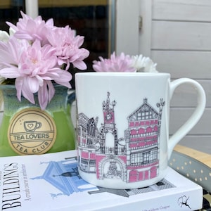 May include: White ceramic mug featuring a black and pink architectural design. The mug is sitting on a book titled "To Read Buildings". A vase of pink flowers and a green jar with "Tea Lovers Tea Club" are in the background.