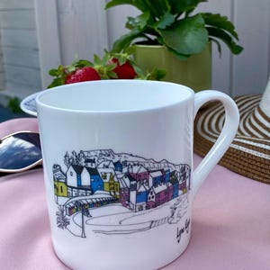Op de afbeelding: Witte keramische mok met een kleurrijke illustratie van een kustplaats, met gebouwen in blauw, geel, roze en paars. De mok heeft een gebogen handvat en de tekst "Lyme Regis" staat op de zijkant.