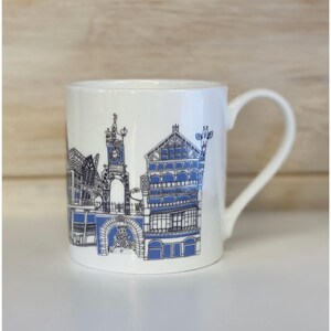 May include: White ceramic mug featuring a blue and black illustration of a cityscape. The design includes buildings, a clock tower, and architectural details. The mug has a curved handle and is set against a neutral background.
