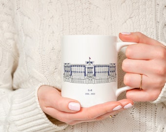 Puede incluir: Una taza de cerámica blanca con un dibujo lineal detallado en azul y gris del Palacio de Buckingham. Debajo de la ilustración, el texto dice E II R y los años 1926-2022 en una fuente serif sencilla. La taza tiene un asa estándar.