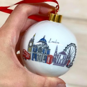 May include: A white spherical Christmas ornament featuring a colorful illustration of London landmarks including Big Ben, St. Paul's Cathedral, a red double-decker bus, a red telephone box, and the London Eye, with the word London written in script.