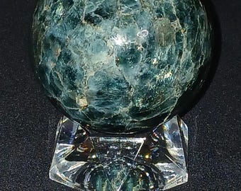 May include: A polished, spherical gemstone in shades of teal and green, resting on a clear, faceted crystal stand. The sphere's surface has a textured, marbled appearance. The stand is a geometric shape, reflecting light.
