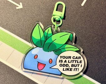Puede incluir: Un llavero acrílico transparente con un personaje de dibujos animados azul con hojas verdes y ojos rojos. El personaje tiene un bocadillo que dice: "¡TU COCHE ES UN POCO RARO, PERO ME GUSTA!" El llavero tiene un cierre verde.