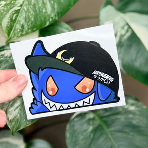 May include: A sticker featuring a blue cartoon character wearing a black baseball cap with a crescent moon. The character has orange eyes and a wide, toothy grin. The text "NATSUKASHII" and Japanese characters are on the cap.