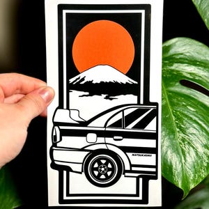 Puede incluir: Adhesivo en blanco y negro con un diseño de coche, una montaña y un sol naranja. El adhesivo tiene el texto "NATSUKASHII" en el lateral. El diseño está enmarcado en un borde negro.