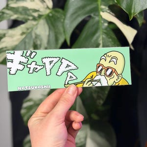 May include: A rectangular sticker with a light green background featuring a cartoon character with a bald head, white eyebrows, and orange sunglasses. The character holds a pipe, and white Japanese text appears as smoke. The word NATSUKASHII is printed in white.