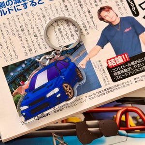 Peut inclure: Porte-clés en forme de voiture bleue avec des roues dorées. Le porte-clés est posé sur un magazine japonais avec du texte et une photo d'une personne. Le porte-clés porte le mot "Option".