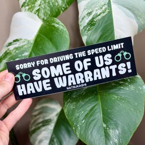 May include: A black bumper sticker with white text that reads "SORRY FOR DRIVING THE SPEED LIMIT SOME OF US HAVE WARRANTS!" with green handcuffs. The sticker is held in front of a variegated green and white plant.