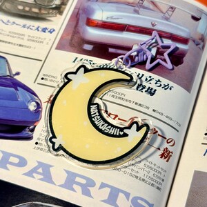 May include: A clear acrylic keychain featuring a yellow crescent moon with the text "NATSUKASHII" and white stars. A purple star-shaped charm and a silver chain are attached. The background is a magazine page with Japanese text and car images.