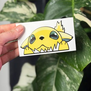 Peut inclure: Un autocollant rectangulaire présentant une illustration de dessin animé d'une créature jaune avec de grands yeux sombres, des oreilles pointues et une petite touffe en forme de flamme sur la tête, dépassant d'un bord blanc avec ses pattes.