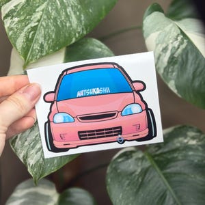 May include: A pink car sticker with a blue windshield and the text "NATSUKASHII". The sticker features a cartoon-style illustration of a car, with black tires and a silver detail on the front bumper. The sticker is held in front of green and white leaves.