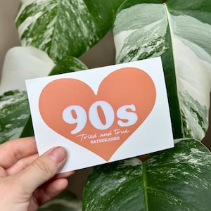 May include: A white sticker with an orange heart shape. The heart contains the word "90s" in white, with "Tried and True" and "NATSUKASHII" written below. The sticker is held in front of a background of green and white variegated plant leaves.