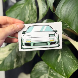 May include: A rectangular sticker featuring a white sports car with pop-up headlights, a front bumper with a large air intake, and yellow turn signal lights. The car illustration is rendered in a clean, graphic style with light blue accents.