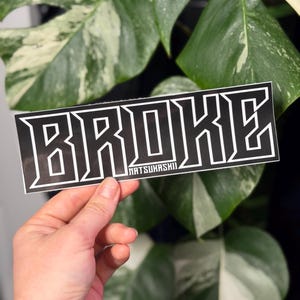 May include: A rectangular sticker featuring the word BROKE in a bold, black, block-style font with a white outline. Below the word, the text NATSUKASHII is printed in a smaller, white, sans-serif font against a black background.