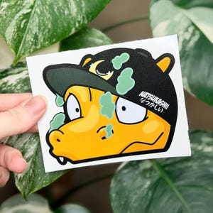 May include: A sticker featuring a cartoon character with a yellow face and a black baseball cap. The cap has green accents and the Japanese word "Natsukashii" printed on it. The character has white eyes and a small mouth.