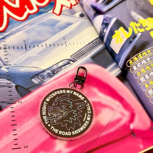 May include: A round keychain with a black wolf design and the text "THE NIGHT WHISPERS MY NAME • THE ROAD ANSWERS MY CALL". The keychain has a black clasp and is on a pink background with a magazine.