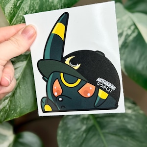May include: A sticker featuring a cartoon character with black and yellow accents, wearing a black baseball cap. The character has orange eyes and a crescent moon design. The text "NATSUKASHII" is printed on the cap.