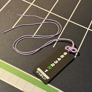 May include: A light purple cord with a decorative knot and a black tag. The tag has white numbers, the word "Natsukashiii" and Japanese text. The background is a black surface with white lines and a green stripe.