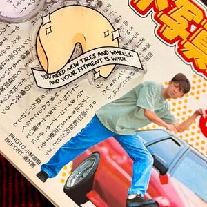 May include: A clear acrylic keychain shaped like a fortune cookie with the text "YOU NEED NEW TIRES AND WHEELS, AND YOUR FITMENT IS WACK." The keychain hangs on a silver chain. The background is a magazine.