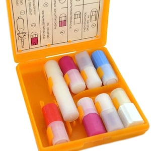 Op de afbeelding: Een oranje plastic koffer met een scharnierend deksel, met daarin acht kleine, witte, cilindrische buizen met gekleurde dopjes. Het deksel heeft een label met instructies in het Russisch, waaronder de woorden "принимать по инструкции" (neem volgens de instructies).