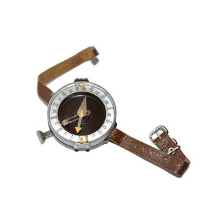 May include: A vintage compass with a dark brown face, a silver metal casing, and a brown leather strap. The compass needle points towards the direction. The leather strap has a buckle. The compass is designed for navigation.