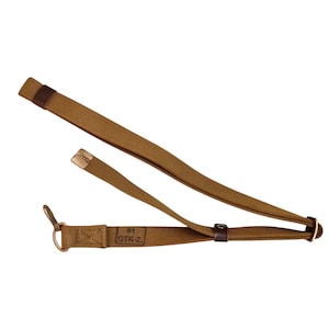 Puede incluir: Una correa de lona color canela con una anilla en D de metal en un extremo, una hebilla deslizante de metal y una trabilla de cuero marrón oscuro. La tela tiene estampado el texto 91 OTK-2 en un recuadro. La correa está doblada e incluye puntas metálicas.