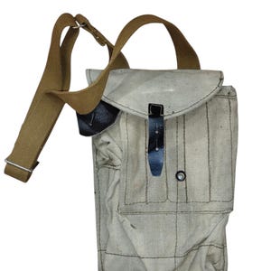 Op de afbeelding: Een lichtgrijze canvas schoudertas met een verstelbare beige riem, een bovenflap met een zwarte leren sluitingsriem en een voorvak dat is vastgezet met een metalen drukknoop. De tas is voorzien van zichtbare stiksels en een rechthoekige vorm.