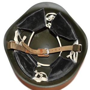 May include: An olive green military helmet, viewed from the inside. It features black leather padding, a brown leather chin strap with a silver buckle, and white cord suspension. The helmet's interior is visible, showcasing its construction.