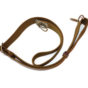 Può includere: Un cinturino in tela marrone con elementi di fissaggio in metallo, tra cui una clip e un gancio. Il cinturino sembra essere per un fucile o altre attrezzature di tipo militare. Il cinturino è realizzato in un materiale tessuto resistente.