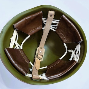 May include: An olive green military helmet interior featuring a brown leather liner system. The liner is suspended by white cord lacing attached to the shell. A tan leather chin strap with a metal buckle and rivets is centered across the opening.