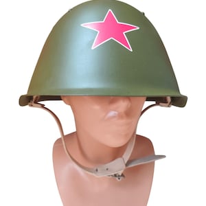 May include: An olive green military helmet with a red star emblem. The helmet has a chin strap and is displayed on a mannequin head. The helmet is a vintage style.