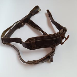 May include: A brown leather strap with metal buckles and stitching. The strap is designed to be worn over the shoulder and has a wide, padded section for comfort.