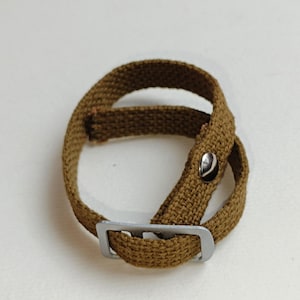 以下が含まれることがあります： 金属製のバックルとボタンが付いた茶色のキャンバスストラップ。ストラップは半分に折り畳まれ、バックルは閉じられています。