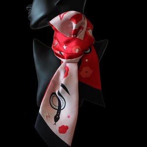 May include: A silk scarf in red and pink, patterned with lips, stars, and a snake motif. The scarf is displayed on a black mannequin head. The scarf has a black border. The design is eye-catching.